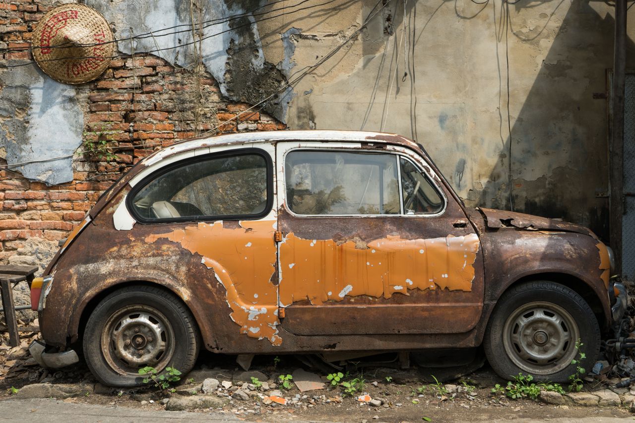 A rusty vintage car parked beside a cracked brick wall in Bangkok, showcasing urban decay.